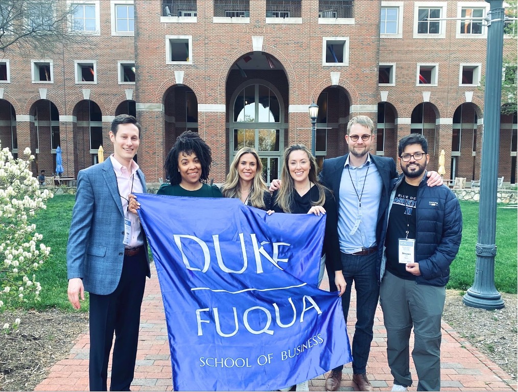 A team of six MBA students in professional outfits stand outside UNC's business school, smiling proudly and holding a Duke Fuqua blue square flag. From left to right: Kyle Ritter, Ayana Ferguson, Lizzy Isgar, Natalie Behan, Mark Dryden, and Shayon Saleh.
